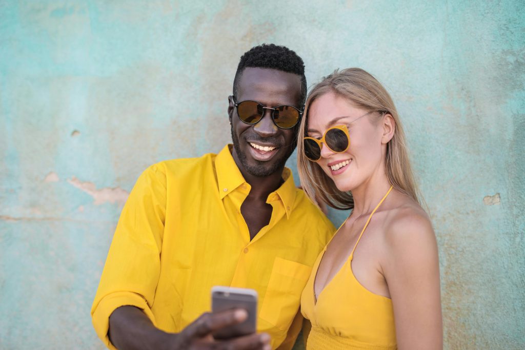 Photo of Smiling Man in Yellow Shirt Standing Beside Smiling Woman in Yellow Spaghetti Strap Top Looking at a Phone