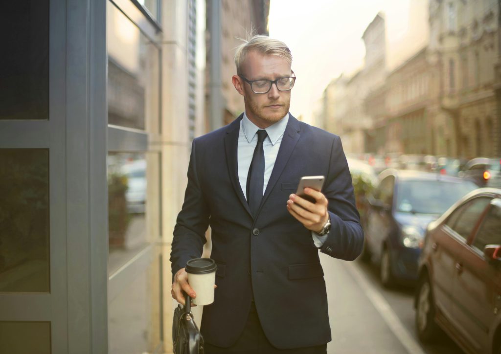 Selective Focus Photo of Walking Man in Black Suit Carrying a To Go Cup and Briefcase While Using His Phone