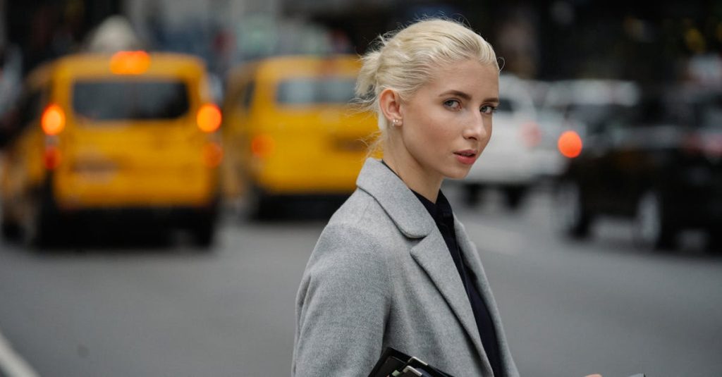 Side view focused young female in formal wear with paper folder and smartphone standing on busy street and looking at camera