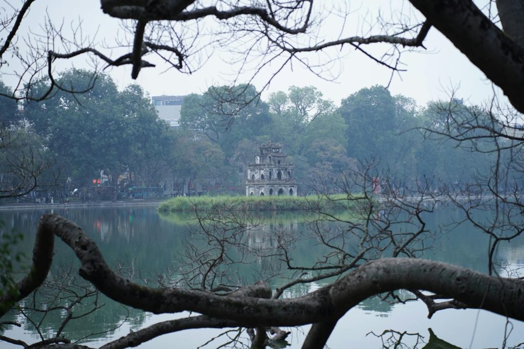 Peaceful view of Turtle Tower in Hanoi framed by branches over serene Hoàn Kiếm Lake.