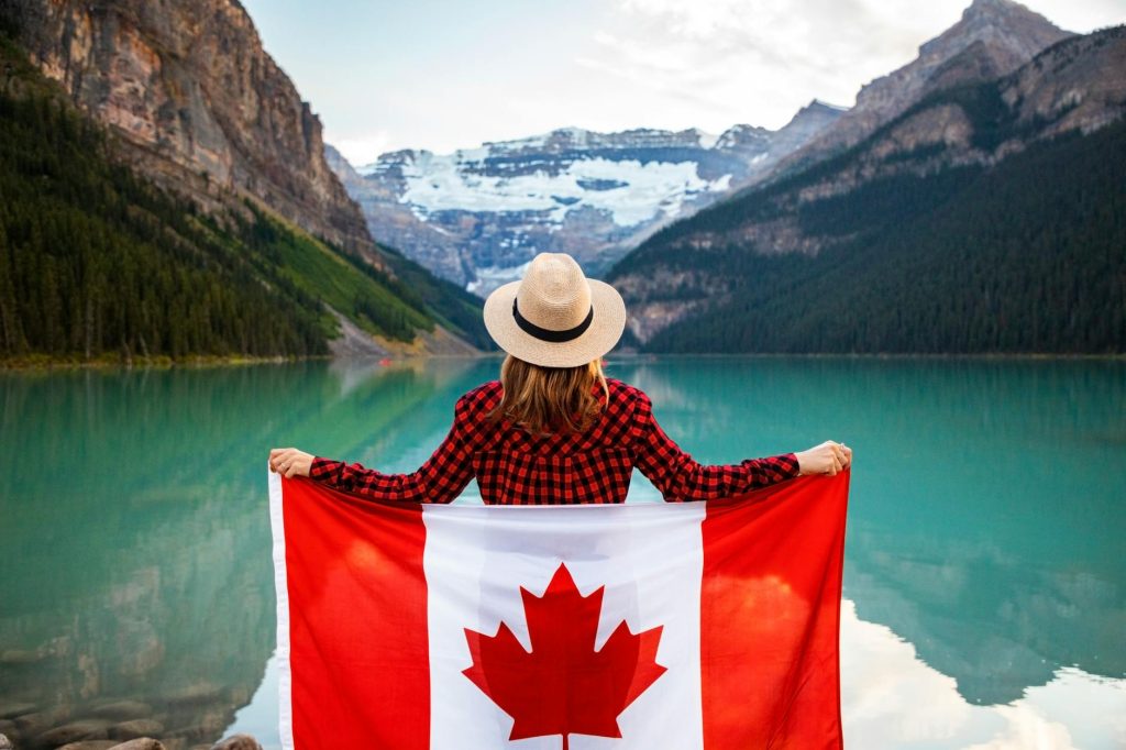 woman wearing red and black checkered dress shirt and beige fedora hat holding canada flag looking at lake